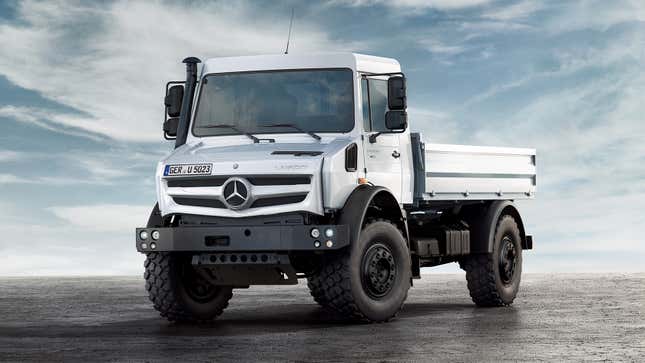 A large silver MBZ Unimog vehicle is parked on a flat, dirt surface in front of a blue sky.